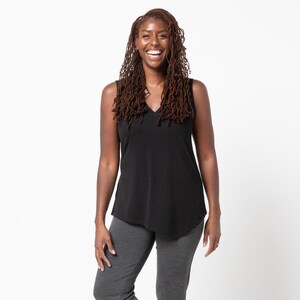 Puede incluir: Una persona lleva una camiseta sin mangas negra con cuello en V y pantalones grises oscuros. Los pantalones están doblados en los tobillos. Lleva tacones negros y sonríe. El fondo es blanco liso.