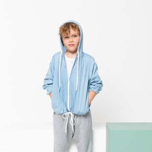 May include: A young person wearing a light blue denim jacket with a hood and a white drawstring. They are wearing gray sweatpants and are standing in front of a light green cube.