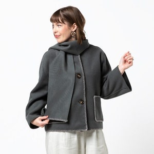 May include: A charcoal gray jacket with a matching scarf, featuring a button closure and white stitching details. The jacket is paired with cream-colored, wide-leg pants and silver sneakers. The outfit is a casual, yet stylish ensemble.