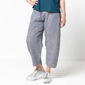 Op de afbeelding: Een vrouw draagt een tealblauw shirt met korte mouwen en een grijze, getailleerde broek. Ze draagt zilveren sneakers.