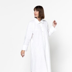 May include: A white linen shirt dress with long sleeves and a button-up front. The dress has a relaxed fit and a slightly curved hemline. The model is wearing silver sneakers.