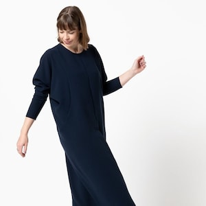 May include: A long, navy blue sweater dress with a relaxed fit and long sleeves. The dress has a single pocket on the left side and a slightly flared hem. The model is wearing the dress with green heeled ankle boots.