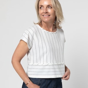 Puede incluir: Una blusa blanca de manga corta con rayas y un dobladillo recortado. La blusa tiene un patrón de rayas finas negras y un detalle ligeramente fruncido en el dobladillo.