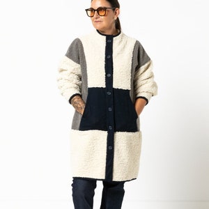 Puede incluir: Una chaqueta larga abotonada con un diseño de patchwork. La chaqueta está hecha de un material suave y peludo y tiene cuello. La chaqueta es en tonos de gris, blanco y negro.