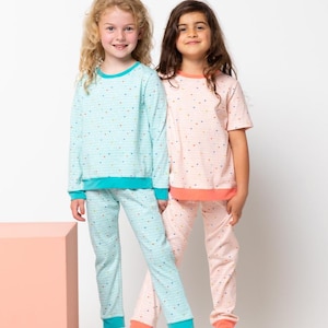 May include: Two children wearing matching pajamas with a colorful confetti print. The pajamas are long-sleeved and have a turquoise and coral trim. The girl on the left is wearing a turquoise top and pants, while the girl on the right is wearing a pink top and pants.