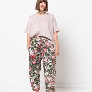 Puede incluir: Una mujer con una blusa rosa claro de manga corta y pantalones con estampado floral. Los pantalones tienen un fondo verde oscuro con grandes rosas rojas, rosas y blancas. También lleva zapatillas de deporte de color rosa claro.