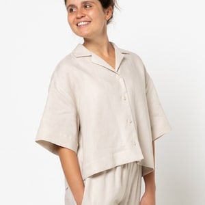 May include: A beige linen shirt and pants set. The shirt has a collared neckline, short sleeves, and a button-up front. The pants have a relaxed fit and a straight leg.