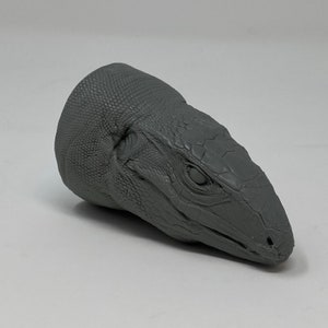 May include: A grey 3D printed model of a lizard head. The model is detailed with scales and a textured surface.