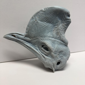 Puede incluir: Un busto esculpido en gris claro de una cabeza de casuario en perfil. La escultura presenta un casco texturizado prominente en la parte superior de la cabeza, un pico afilado y texturas de piel detalladas alrededor del ojo y las fosas nasales.