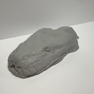 May include: A gray, three-dimensional sculpture of a snake head featuring detailed, textured scales across the surface. The object is shaped like a realistic serpent head with defined eye sockets and a snout, presented in a solid, matte gray finish.