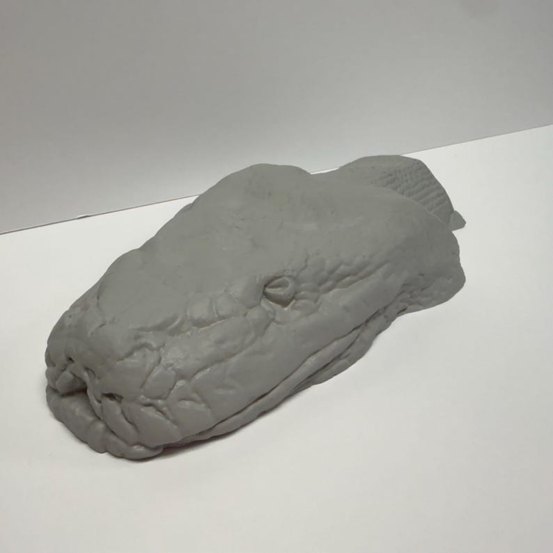 May include: A gray, three-dimensional sculpture of a snake head featuring detailed, textured scales across the surface. The object is shaped like a realistic serpent head with defined eye sockets and a snout, presented in a solid, matte gray finish.