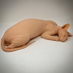 Könnte beinhalten: Figur einer haarlose Sphynx-Katze in Schlafposition. Die Katze hat eine hellpfirsichfarbene Haut, geschlossene Augen und eingeklappte Pfoten. Der Schwanz ist eingerollt und die Ohren aufgestellt. Die Figur liegt auf einer weißen Oberfläche.