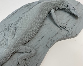 Peut inclure: Modèle 3D imprimé en gris d'un lézard sur un fond gris texturé. Le lézard est posé la tête tournée sur le côté et les pattes tendues.
