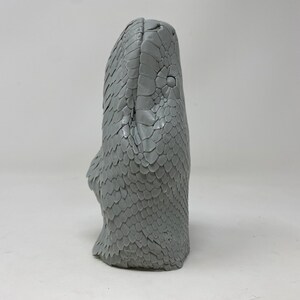 May include: A gray sculpture of a snake's head, showcasing detailed scales and a textured surface. The snake's head is upright, with its eyes and facial features clearly defined. The sculpture is set against a plain white background.