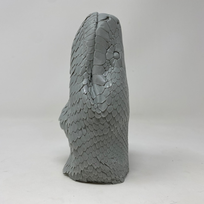 May include: A gray sculpture of a snake's head, showcasing detailed scales and a textured surface. The snake's head is upright, with its eyes and facial features clearly defined. The sculpture is set against a plain white background.