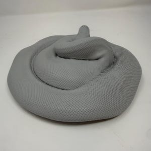 Pu&ograve; includere: Una scultura grigia solida di un serpente avvolto su se stesso con un motivo a squame dettagliato e testurizzato che copre l'intero corpo. Il serpente &egrave; posizionato in un cerchio stretto con la testa leggermente sollevata sulla parte centrale.