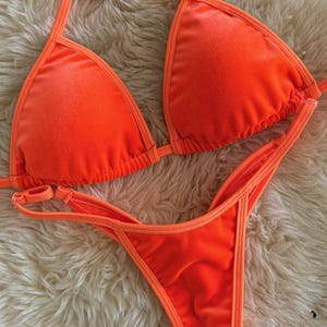 May include: Bright orange bikini set. The top is a triangle style with thin straps, and the bottom is a thong style. The edges are trimmed with a lighter orange. The set is displayed on a fluffy, white surface.