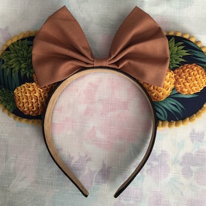 May include: Brown fabric bow headband with pineapple print fabric ear covers. The ear covers have a yellow trim.