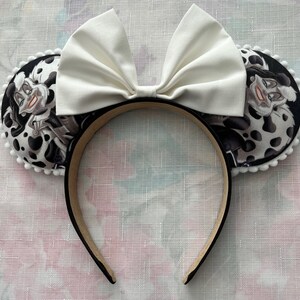 May include: A pair of black and white fabric Minnie Mouse ears with a white bow. The ears feature a Dalmatian print with a cartoon image of Cruella de Vil.