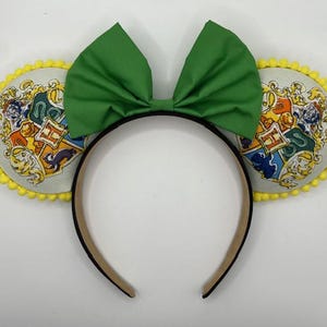 May include: A headband with two round, cream-colored ears featuring a colorful crest design and yellow pom-pom trim. A large green bow sits atop the black headband. The crest design includes a shield with various symbols.
