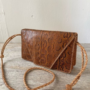 Può includere: Una borsa a tracolla in pelle di serpente marrone con una tracolla in pelle intrecciata. La borsa ha una chiusura a lembo e un design geometrico.