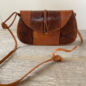 May include: A brown leather purse with a snakeskin flap and a long strap. The purse has a small, round button closure.