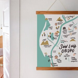 May include: A hand-drawn map of San Luis Obispo, California, with a teal background and brown lines. The map highlights various landmarks, including Morro Rock, Morro Bay, Avila Beach, Pismo Beach, and Cal Poly. The map is framed in a light wood frame.
