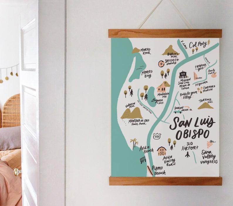 May include: A hand-drawn map of San Luis Obispo, California, with a teal background and brown lines. The map highlights various landmarks, including Morro Rock, Morro Bay, Avila Beach, Pismo Beach, and Cal Poly. The map is framed in a light wood frame.