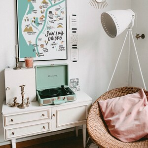 May include: A white dresser with three drawers and gold knobs. A mint green record player is on top of the dresser. A wicker chair with a pink pillow is in the background. A map of San Luis Obispo is hanging on the wall. The map is in a black frame and has a light blue background with white and black text. The text on the map includes "San Luis Obispo", "SLO Airport", "Edna Valley", and "Pismo Beach".