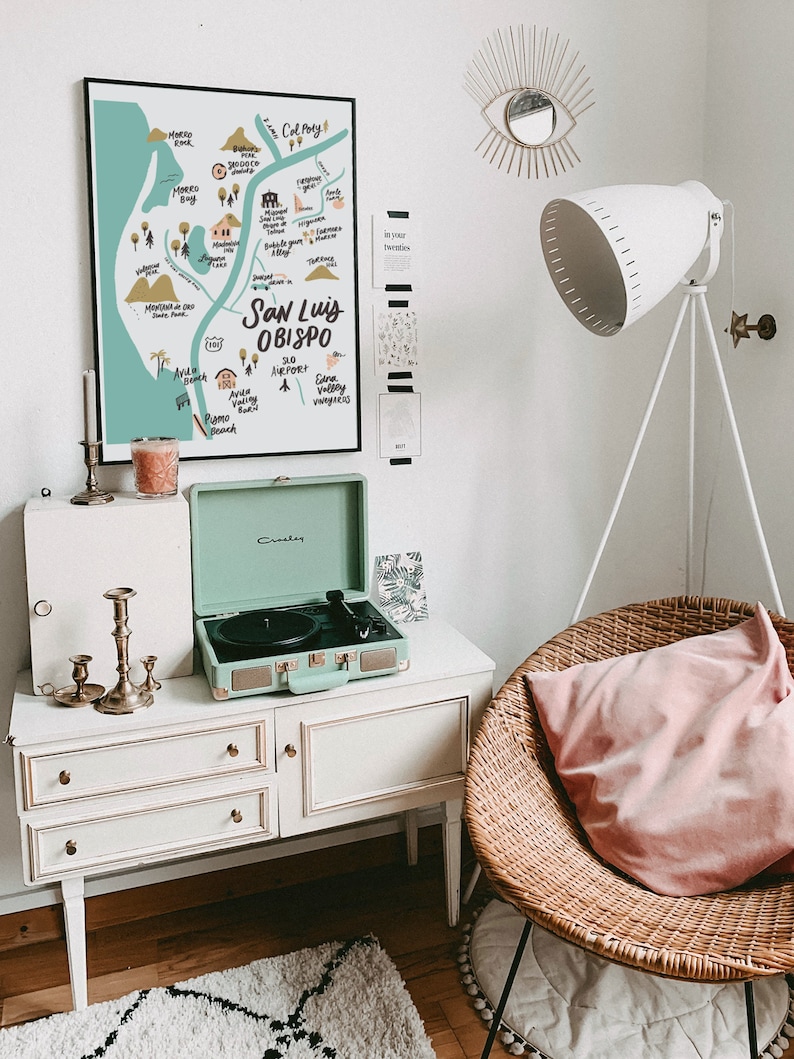 May include: A white dresser with three drawers and gold knobs. A mint green record player is on top of the dresser. A wicker chair with a pink pillow is in the background. A map of San Luis Obispo is hanging on the wall. The map is in a black frame and has a light blue background with white and black text. The text on the map includes "San Luis Obispo", "SLO Airport", "Edna Valley", and "Pismo Beach".