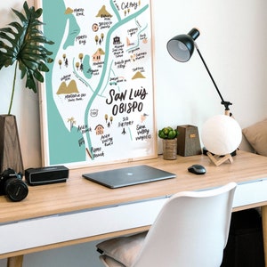 May include: A framed illustration of a map of San Luis Obispo, California, with hand-drawn details and landmarks. The map is on a light blue background with brown and yellow accents. The map is hanging on a white wall above a wooden desk with a laptop, a lamp, and a white chair.