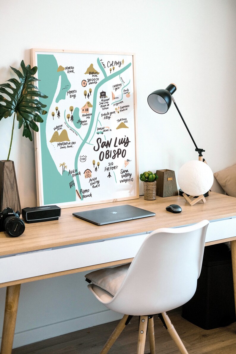 May include: A framed illustration of a map of San Luis Obispo, California, with hand-drawn details and landmarks. The map is on a light blue background with brown and yellow accents. The map is hanging on a white wall above a wooden desk with a laptop, a lamp, and a white chair.