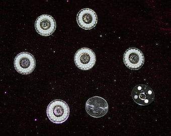 May include: Seven white and silver round embellishments with a pearl-like border. The embellishments are on a dark burgundy fabric background.