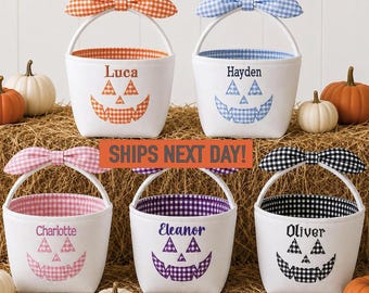 May include: Five white fabric baskets with gingham-patterned bows and jack-o'-lantern faces. Each basket features a personalized name and matching gingham interior lining in orange, blue, pink, purple, or black. Small pumpkins are placed around.