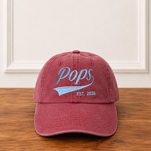 May include: A maroon baseball cap featuring the word Pops embroidered in light blue script font with a light blue swoosh underneath. The text EST. 2025 is embroidered in light blue to the right of the swoosh. The cap has a curved brim.