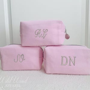 May include: Three light pink cosmetic bags with a textured waffle weave pattern. Each bag has a silver zipper and embroidered initials: RL, SO, and DN. The bags are arranged in a stacked formation.