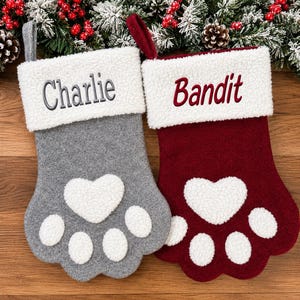 May include: Two paw-shaped Christmas stockings with white sherpa cuffs and white sherpa paw pads. The gray stocking features the embroidered name Charlie, and the burgundy stocking features the embroidered name Bandit. Both have hanging loops.