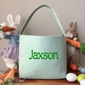 May include: A green and white striped Easter basket with the name "Jaxson" embroidered in green. The basket has a matching green handle. Two stuffed bunny rabbits and carrots are in the background. The Wild Wind Apparel logo is in the lower left corner.