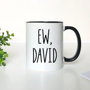 May include: White ceramic coffee mug with a black rim and handle. The mug has black text that reads "EW, DAVID".