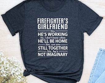May include: Dark gray t-shirt with white text that reads "FIREFIGHTER'S GIRLFRIEND" followed by a series of statements: "Yes, HE'S WORKING", "No, I don't know when HE'LL BE HOME", "Yes, we are STILL TOGETHER", and "No, he's NOT IMAGINARY".