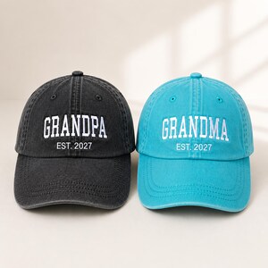 May include: A black baseball cap with white embroidered text reading GRANDPA EST. 2027 and a turquoise baseball cap with white embroidered text reading GRANDMA EST. 2027. Both caps feature a six-panel construction with a curved brim and top button.