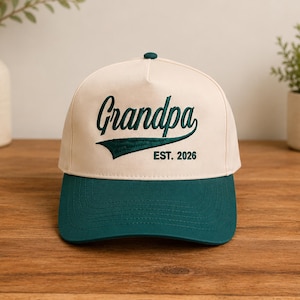 May include: A baseball cap with a cream-colored crown and a dark green brim. The word "Grandpa" is embroidered in dark green script across the front, with "EST. 2026" below it. The cap is sitting on a wooden surface.