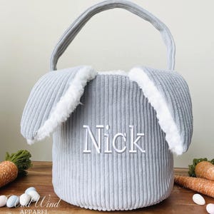 May include: Gray corduroy Easter basket with bunny ears and white trim. The basket has a handle and is personalized with the name "Nick".