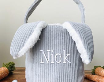 May include: A gray corduroy Easter basket shaped like a bunny, with long ears lined with white faux fur. The name "Nick" is embroidered on the front. Carrots and Easter eggs are in the background.