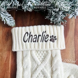 May include: White cable knit stocking with the name "Charlie" and a paw print embroidered in brown on the cuff.