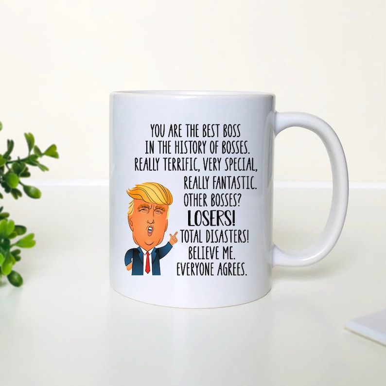 Pode incluir: Caneca de cer&acirc;mica branca com uma imagem de desenho animado de Donald Trump. A caneca tem texto que diz "Voc&ecirc; &eacute; o melhor chefe na hist&oacute;ria dos chefes. Realmente &oacute;timo, muito especial, realmente fant&aacute;stico. Outros chefes? Perdedores! Desastres totais! Acredite em mim. Todos concordam."