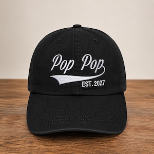 May include: A black baseball cap featuring white embroidered text that reads Pop Pop with a swoosh underneath and the text EST. 2027 below it. The cap has a structured crown, a curved brim, and a button on top, showcasing a classic six-panel design.