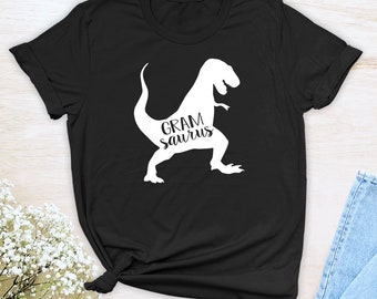 Pode incluir: Camiseta preta com uma silhueta branca de um dinossauro e o texto "GRAM saurus" impresso na frente.