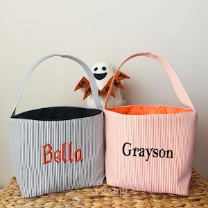 May include: Two striped Halloween treat bags. One is navy and white with orange embroidery that says "Bella". The other is peach and white with black embroidery that says "Grayson". Both have handles.