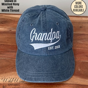 May include: A blue washed cotton baseball cap with white embroidery that reads "Grandpa EST. 2023".